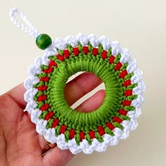 a hand holding a green and red crochet christmas wreath ornament on a white background