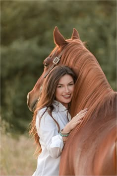 a beautiful young woman standing next to a brown horse with her head on the back of it