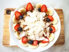 a white bowl filled with bananas, strawberries and granola on top of a wooden cutting board