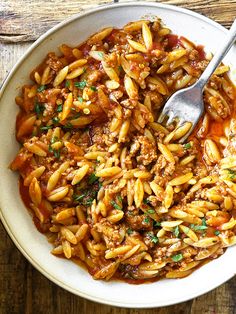 a white bowl filled with pasta and meat on top of a wooden table next to a fork