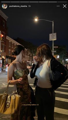 two women standing next to each other on a street corner at night with one holding a purse