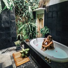 a woman sitting in a bathtub next to a wooden table and potted plants