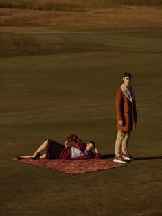 two people are laying on a blanket in the middle of a golf course while one person is standing