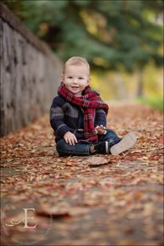 a little boy sitting on the ground with leaves all over him and wearing a scarf around his neck