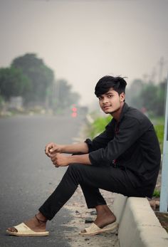 a young man sitting on the side of a road next to a street sign with trees in the background