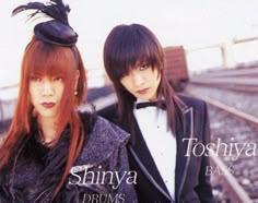 two young women standing next to each other in front of a train track with the words shinya drums on it