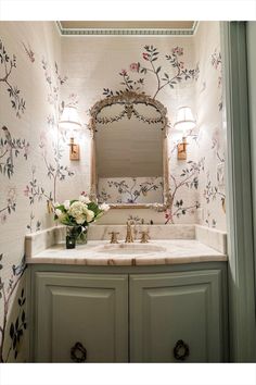 a bathroom with floral wallpaper and blue cabinetry, gold mirror above the sink