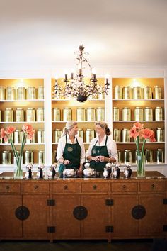 two women in aprons standing behind a counter with vases and bottles on it