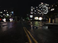 an empty city street at night with buildings lit up in the background and rain on the ground