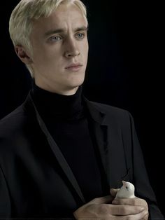 a man in a black suit and turtle neck sweater holding a small white object with one hand