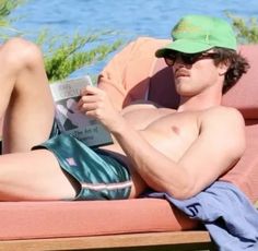 a man laying on top of a pink couch next to the ocean reading a book