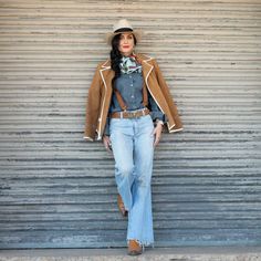 a woman standing in front of a metal wall wearing jeans and a cowboy style hat