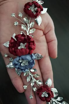 a person's hand holding three red and blue flowers with silver leaves on them