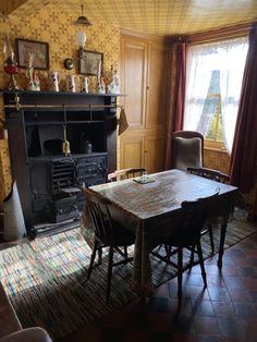 an old fashioned dining room table and chairs in front of a fireplace with ornate wallpaper