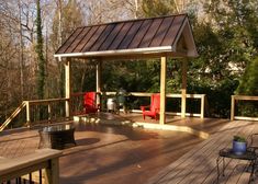 a wooden deck with red chairs and a grill on it's side, surrounded by trees