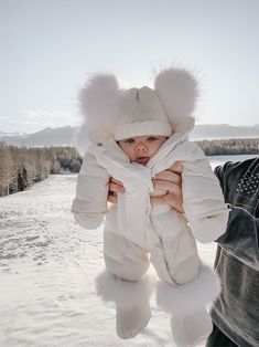 a man holding a baby wearing a white hat and coat in the snow with mountains in the background