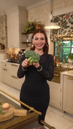 a woman standing in a kitchen holding a piece of broccoli and smiling at the camera