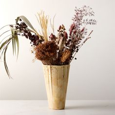 an arrangement of dried flowers in a vase on a white countertop with a planter behind it