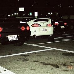 two cars are parked in a parking lot at night, one is white and the other is black