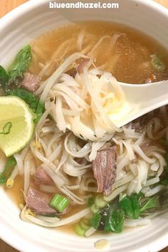 a white bowl filled with noodles and meat, garnished with green onions next to a lemon wedge