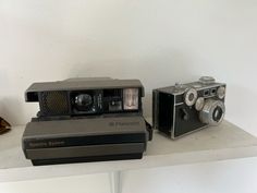 two old cameras sitting on top of a shelf next to each other, one is black and the other is gray