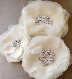 three white flower brooches with crystal centers on a gray background, set against a light colored backdrop