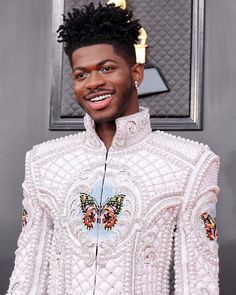 a man with an afro standing in front of a gray wall wearing a white jacket