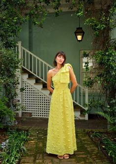 a woman in a yellow dress standing on a brick walkway next to some bushes and trees