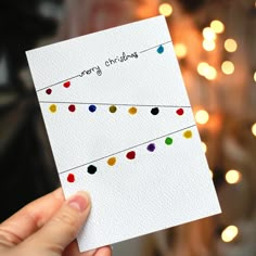 a hand holding up a christmas card in front of a tree with lights on it