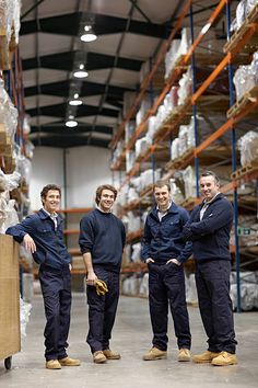 three men in blue work clothes standing next to each other with boxes on the shelves behind them