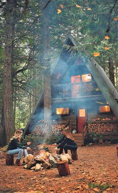 two people sitting around a campfire in front of a log cabin surrounded by trees