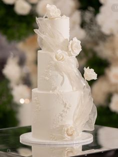 a three tiered white wedding cake with flowers on the top and bottom, sitting on a glass table