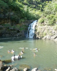 many people are swimming in the water near a waterfall and some rocks, while others stand around