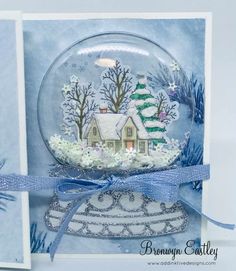 a snow globe with a house in it and blue ribbon around the edge, sitting on top of a card