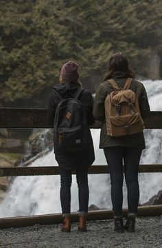 two people standing next to each other with backpacks on looking at a water fall