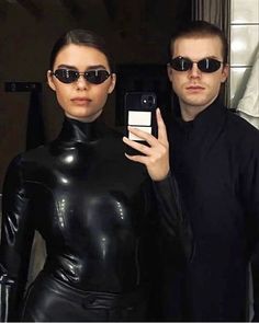 a man and woman in black catsuits taking a selfie with their cell phones