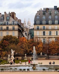 people sitting on benches near a fountain in the middle of a park with trees and buildings behind them