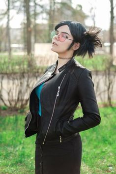 a woman with black hair and glasses is standing in the grass wearing a black jacket