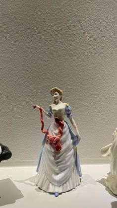 three porcelain figurines are on display in a museum case, one is wearing a white dress and the other has a red ribbon