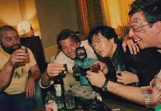 four men are drinking and laughing while sitting at a table in a room with other people