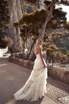 a woman in a white dress is standing near some trees and rocks with her back to the camera