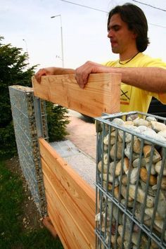 a man standing next to a pile of rocks in a metal basket on top of a wooden fence