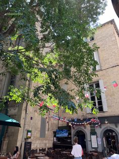 people are sitting at tables under the shade of a tree in an old city square