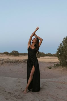 a woman in a black dress is standing on the sand with her arms raised up