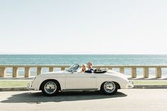 a man and woman are sitting in an old white convertible car by the ocean,