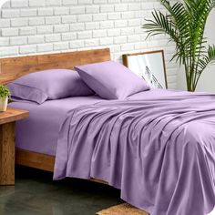 a bed with purple sheets and pillows in a room next to a potted plant