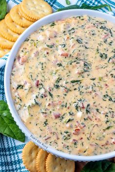 a white bowl filled with cheese and spinach dip surrounded by crackers on a blue checkered table cloth