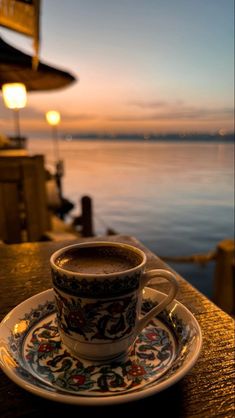 a cup of coffee sitting on top of a saucer next to the ocean at sunset