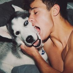 a man laying on top of a bed with his mouth open next to a husky dog