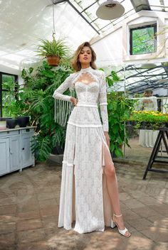 a woman standing in a greenhouse wearing a white dress with long sleeves and high slit